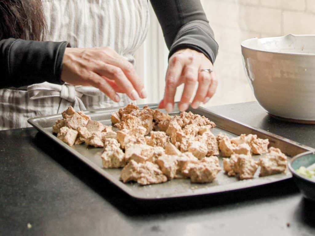 A person wearing a striped apron arranges pieces of textured, tofu on a baking tray. There is a large white bowl nearby. The scene suggests preparation for baking or roasting.